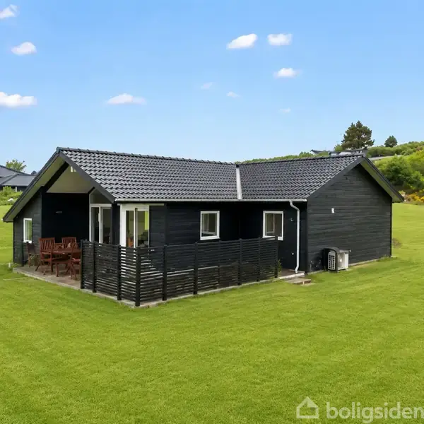 Sort sommerhus med stakit rundt terrassen, der har havemøbler. Huset ligger på en stor græsplæne med spredte buske og træer i baggrunden under en blå himmel.