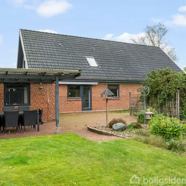 Rødt murstenshus med sort tag, terrasse med havebord og stole under pergola. Omgivet af have med græsplæne, planter og en lille stenbelagt sti i en rolig boligområde.