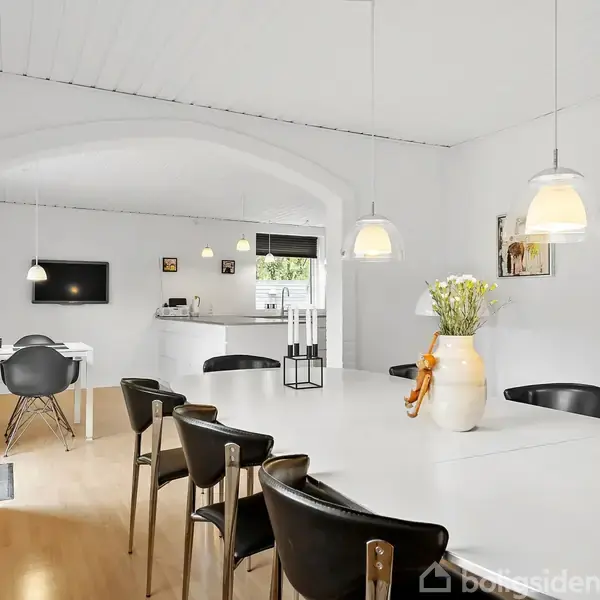 En spisestue med et langt, hvidt bord og sorte stole er placeret centralt under hængende lamper. I baggrunden ses et køkken. Malerier hænger på de hvide vægge.