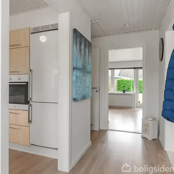 Køkken med hvid ovn og køleskab integreret i træskabe. En blå jakke hænger på væggen. Gennem en åben dør ses en stue med store vinduer og udsigt til grønne buske.