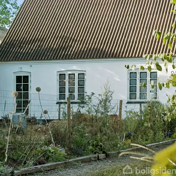 Et hvidt hus med mørkt tag står i en have omgivet af et lavt hegn. Foran huset er der forskellige planter, og omgivelserne er frodige med grønne træer.
