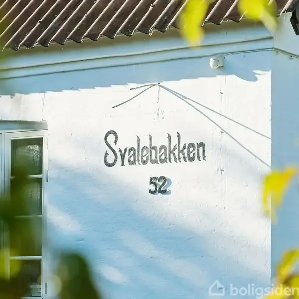 Hvid husvæg med teksten "Svalebakken 52". En del af et vindue ses til venstre. Forgrunden viser uklare, grønne blade, som skaber en indrammende effekt.