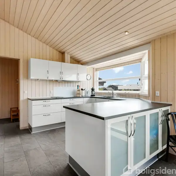 Et moderne køkken med hvide skabe og sorte bordplader. Rummet er lyst med trægulv, træbeklædte vægge og et stort vindue med udsigt til blå himmel og hus. En sort stol står ved bordet.