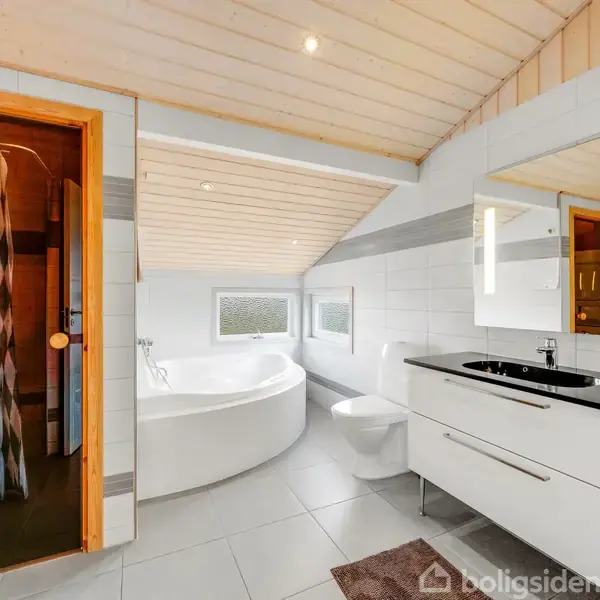 Træbeklædt badeværelse med spabad i hjørnet, sauna til venstre bag en glasdør, hvidt møblement med sort vask til højre, flisegulv og skråt loft med indbyggede spots.