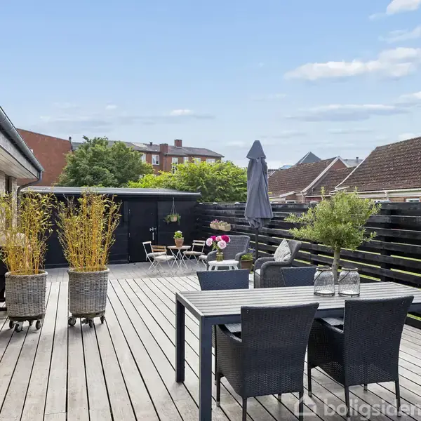 Terrasse med træbord og stole omgivet af grønne planter i krukker. En parasol står ved siden af bordet. Den solrige, åbne omgivelser inkluderer bygningers tage og en himmel med få skyer.