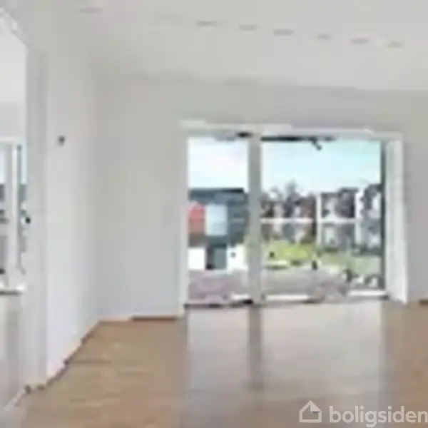 Tomt rum med hvidmalede vægge og trægulv. Store vinduer åbner mod en balkon med udsigt til moderne bygninger og træer i en solrig urban kontekst.