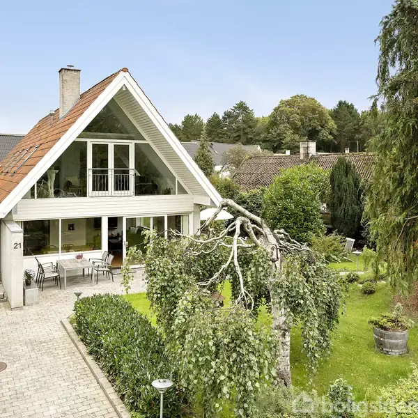 En moderniseret villa med gavlvindue og rød tagstenstag. Omkring huset er en velplejet have med græsplæne og træer. En terrasse foran indgangen har havemøbler. Husnummeret "21" er synligt.