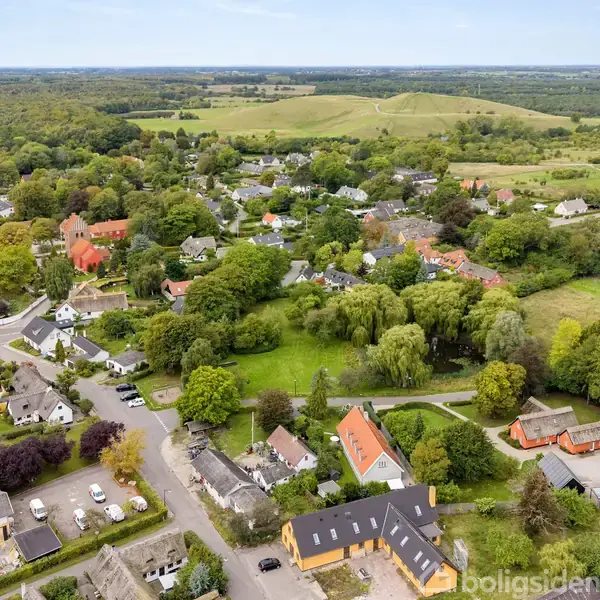 Et landskab med en lille by omgivet af grønne marker og skove. Huse med farverige tage ses blandt træerne, og veje snor sig gennem området.