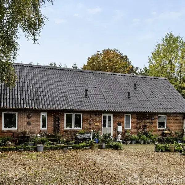 Et rødt murstenshus med et sort tegltag står i en have. Foran huset er der mange blomsterpotter, og huset er omgivet af grønne træer og buske.