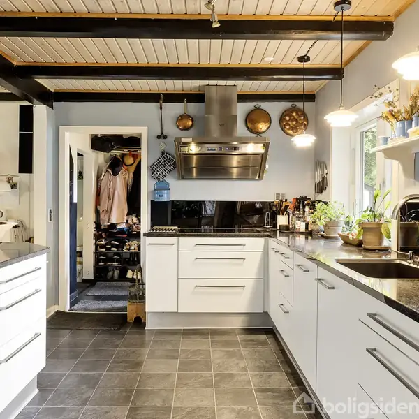 Køkken i moderne stil har hvide skabe og sort bordplade. Det er velorganiseret med hængende gryder, planter og køkkenapparater. Lyse lamper hænger fra loftet, og et vindue giver dagslys.