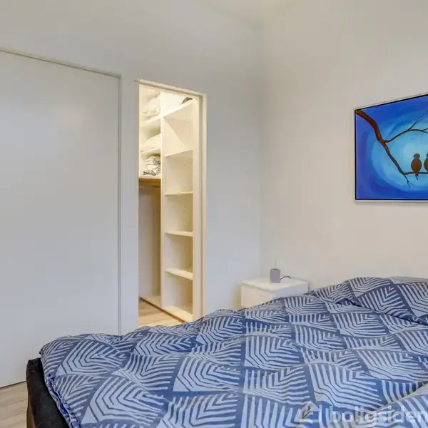 Seng med mønstrede sengetæpper står i soveværelse. I baggrunden ses et walk-in closet med åbne hylder. På væggen hænger et maleri af to fugle på en gren.