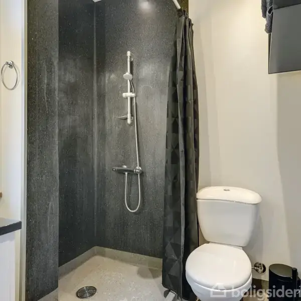 En brusekabine med sort væg og bruserhoved, står ved siden af et toilet i et lyst badeværelse. Dusjforhæng er sort, og der er et håndklædering på væggen.