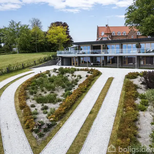 Moderne hus med store glaspartier står stille i en velplejet have med snoede grusstier og blomsterbede. Græs og træer omkranser området, mens en blå himmel ses i baggrunden.