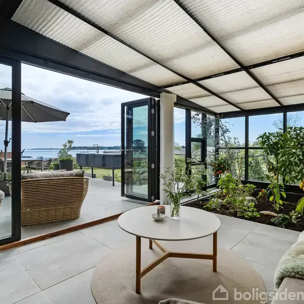 En lys stue med glasvægge åbner ud til en terrasse med havudsigt. I stuen står en rund sofabord med en vase, omgivet af planter og en sofa.