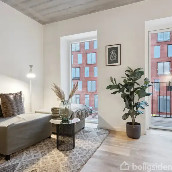 En stue indeholder en grå sofa med puder, en gulvlampe ved siden af. Der er et tæppe på gulvet, et bord med planter, og en stor plante ved glasdøren med udsigt til en murstensbygning udenfor.