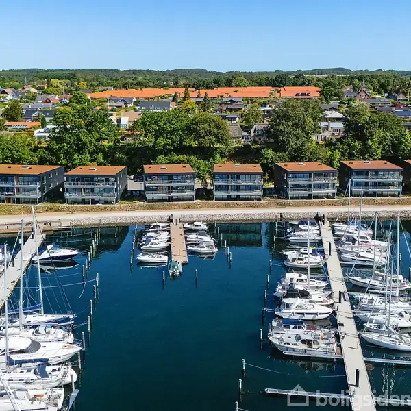 Både ligger til kaj i en moderne marina med rækker af lyse boligbygninger i baggrunden, omgivet af grønne træer og en by i det fjerne.