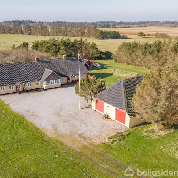 Gårdanlæg med gulmalet bygning og flagstang står stille på en stor grusbelagt gårdsplads omgivet af grønne marker og skov i landlig omgivelser.