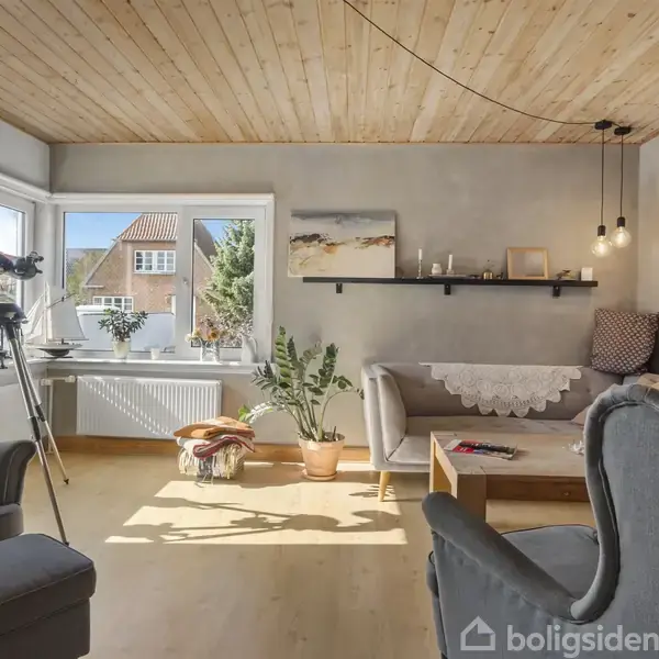 En stue med grå sofa og lænestole omkring et sofabord. Store vinduer lader sollys ind, og en kikkert står ved siden af planter. Væghylder har dekorationer.