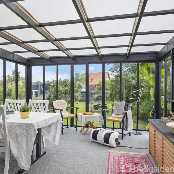 En overdækket terrasse med spisebord dækket af en hvid dug. En hund ligger på gulvet. Store vinduer giver udsigt til en grøn have og røde hustage.