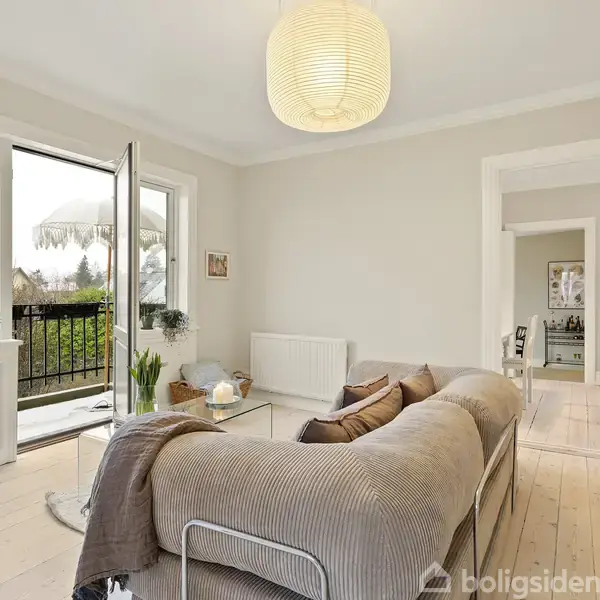 En beige sofa står foran en åben altandør og et sofabord med dekorationer; omgivet af planter i en lys stue med trægulv og en lampe hængende fra loftet.