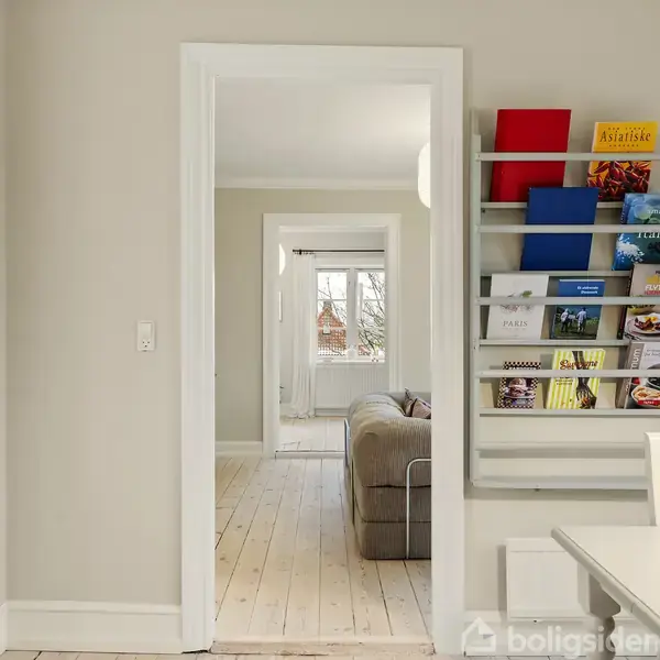 En vægmonteret tidsskriftholder indeholder farverige magasiner. Ved siden af er der en åben døråbning, hvor et sofahjørne er synligt. Trægulve og lyse vægge omgiver området.