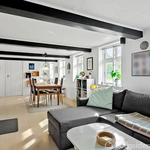 Stor, lys stue med grå sofa og sofabord i forgrunden. I baggrunden er der et spisebord med stole. Rummet har lyse vægge, lyse trægulve og synlige loftsbjælker.