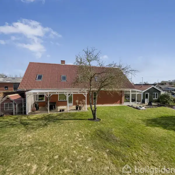 Hus med rødt tag står i en grøn have. Det er omgivet af andre huse og har en træterrasse og et fritstående træ i forgrunden.