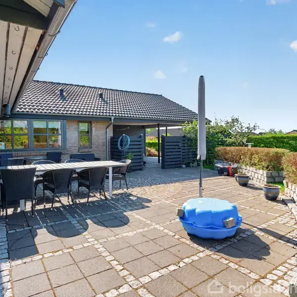 En gårdhave med fliser har et bord og stolegruppe på den ene side. Modstående væg har en stor, åben parasol ved siden af en blå plastiksandkasse. Omkring er der hække og græsplæne.