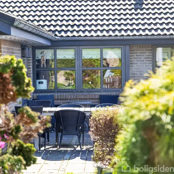 Terrassebord med stole står på flisebelagt gårdhave. Omgivet af grøn beplantning og buske. Murstenshus med store vinduer danner baggrunden. Sollyset skaber en lyst og indbydende atmosfære.