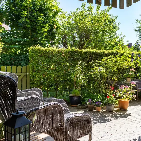Fletstole står på en brolagt terrasse med en lanterne foran. Omgivet af frodige grønne planter og buske, skaber det en hyggelig haveatmosfære.