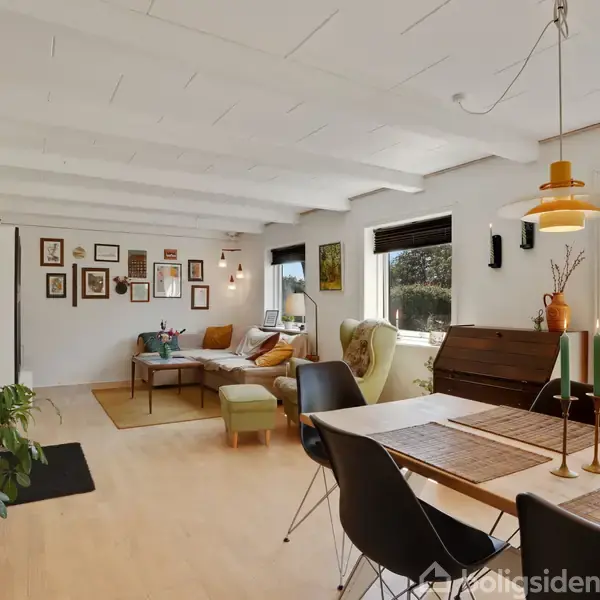 Stue med sofaområde, dækket spisebord og moderne stole. Langs væggen hænger billeder. Store vinduer slipper lys ind og giver udsigt til en grøn have.