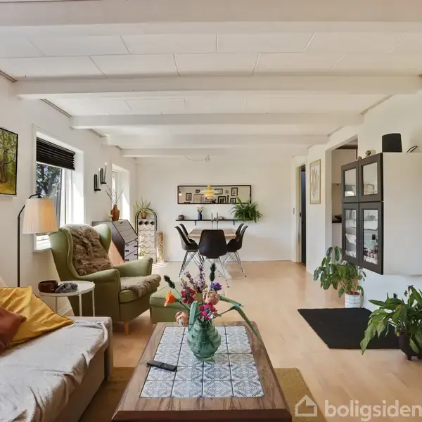 Stue med sofa og lænestol i forgrunden, blomster på sofabordet. Spisebord med stole og planter i baggrunden, omgivet af et lyst rum med hvide vægge og vinduer.