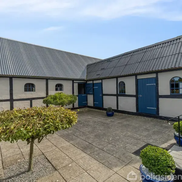 En gårdbygning med hvide vægge og sort bindingsværk står stille. Flisebelagt gårdsplads omgives af trimmede buske og blåmalede døre. Klar himmel i baggrunden.