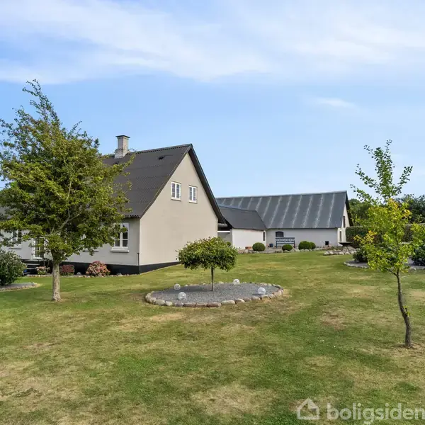 Et beige hus ligger i en velplejet have med grønne plæner og flere små træer. I baggrunden ses en stor, grå lade. Himlen er blå med lette skyer.