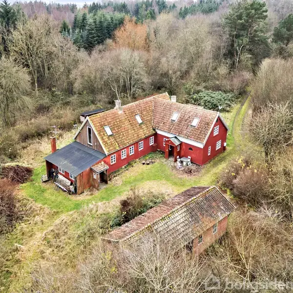 Rødt hus med skråt tag omgivet af skov. Husets ene side har en forstue med sort tag. En mindre bygning ligger i nærheden, alt i et naturligt, skovrigt område.