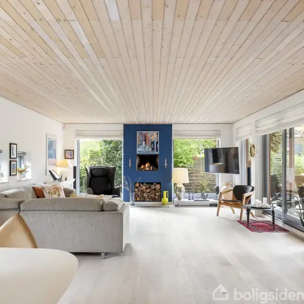 Stue med grå sofa og lænestol står foran en brændeovn. Store vinduer åbner til en grøn have. Lyse trægulve og hvidt loft skaber en lys atmosfære.
