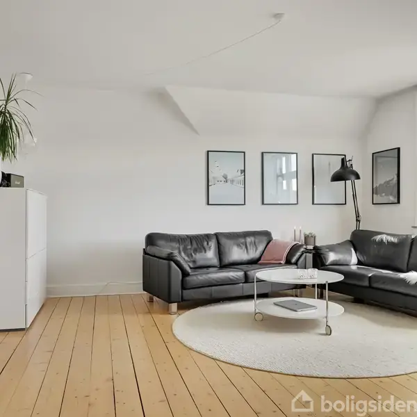 Sort lædersofa står i en stue på et lysebrunt trægulv, flankeret af et rundt, hvidt sofabord og en stående lampe. Væggen har indrammede billeder og et skab med en plante.