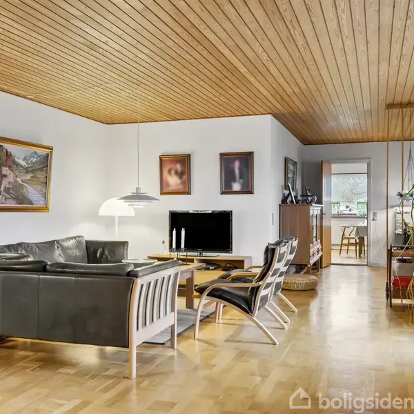 Sort læder sofaarrangement står i stuen med lakeret trægulv og træloft. Malerier hænger på væggen, og store planter sidder langs vinduet. I baggrunden ses en åbning til køkkenet.