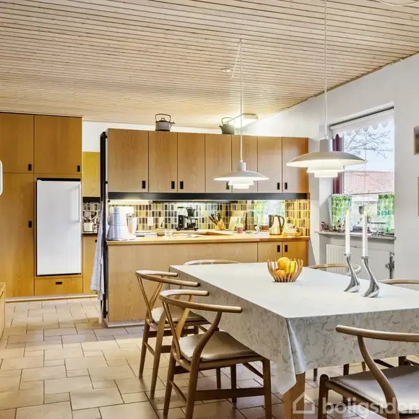 Et køkken med et spisebord dækket af en dug, omgivet af stole, står i forgrunden. I baggrunden ses køkkenskabe, en indbygget ovn og køleskab. Vinduer slipper dagslys ind.