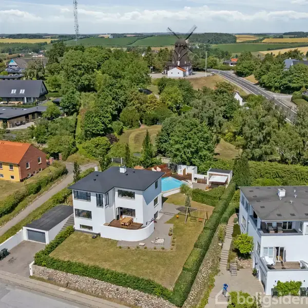 To hvide moderne huse med flade tage ligger på grønne grunde omgivet af buske og små veje. I baggrunden ses en mølle på en bakketop, omringet af frodige træer.