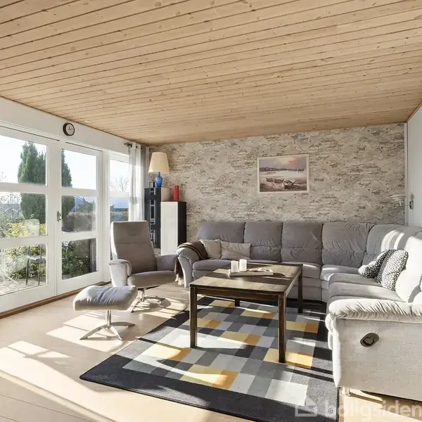 En grå sofa står i en lys stue med trægulv og træloft. Store glasdøre lader sollys strømme ind. Moderne vægtapet og farverigt tæppe giver et hyggeligt præg.