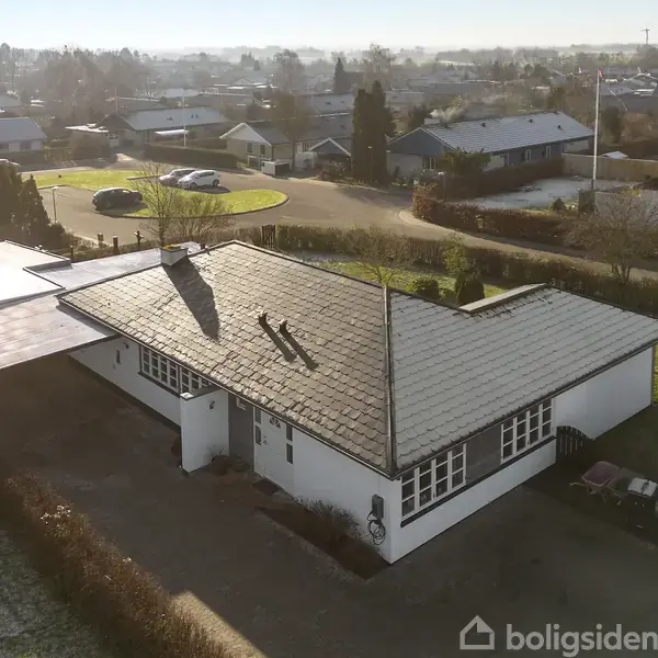 Et hvidt hus med fladt tag står stille i et villakvarter. Omgivet af en have, ses det fra fugleperspektiv på en solrig dag.