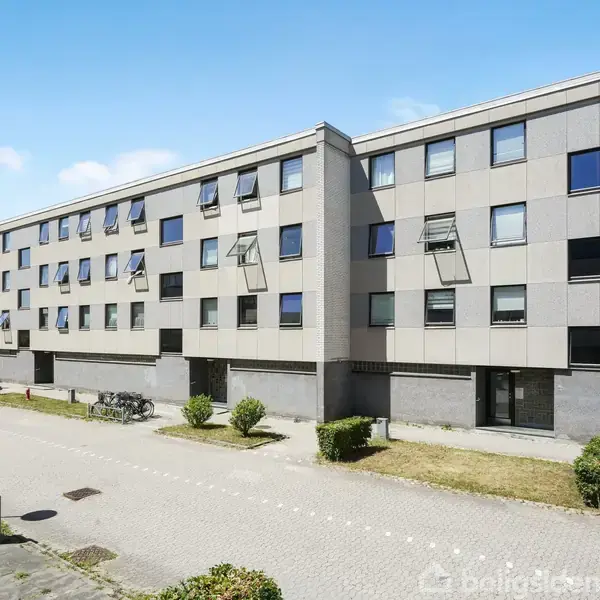 Et langt, gråt boligkompleks med mange vinduer står langs en bred, flisebelagt gangsti omkranset af små buske under en blå himmel.