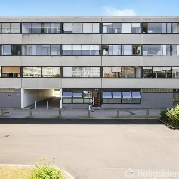En fleretagers bygning med vinduer vises, mens en rød bil holder parkeret i forgrunden. Bygningen er omkranset af asfalt og buske. Der er ingen tekst at transskribere.