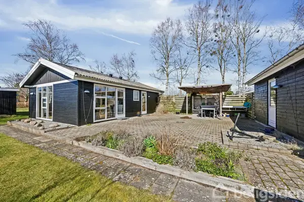 Et moderne sommerhus med glasdøre står stille ved siden af en flisebelagt terrasse og overdækket opholdsområde. Omgivet af et velplejet grønt område og høje, bladløse træer i baggrunden.