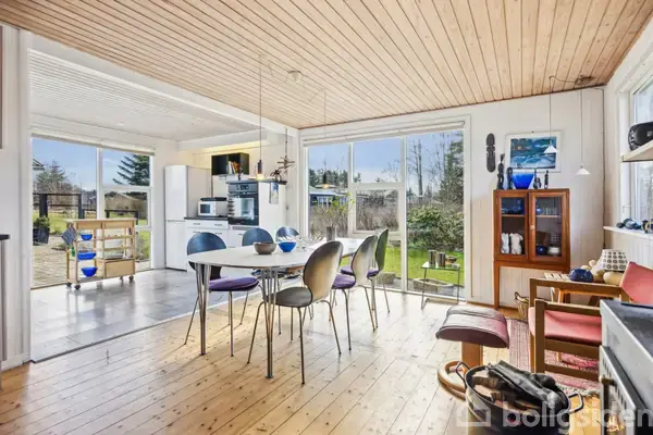 Et lyst spiseområde med et bord og seks stole i et rum med store vinduer, der viser en have udenfor. I baggrunden ses et køkkenområde.