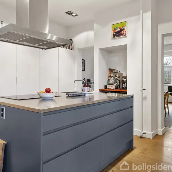 Køkkenø med lys træbordplade står centralt. To tomater er placeret på overfladen. I baggrunden ses hvide skabe, en kaffemaskine og et spisebord nær et vindue.