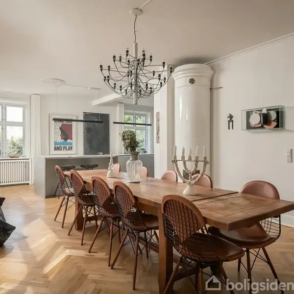 Stor, lys spisestue med lang træbord, omgivet af otte stole. En moderne lysekrone hænger fra loftet. Rummet har parketgulv og hvide vægge, med et vægtæppe og store vinduer.