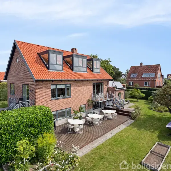 Rødt murstenshus med rødt tegltag, har en stor træterrasse med hvide havemøbler. Omgivet af velplejet have med buske og træer i et parcelhuskvarter.