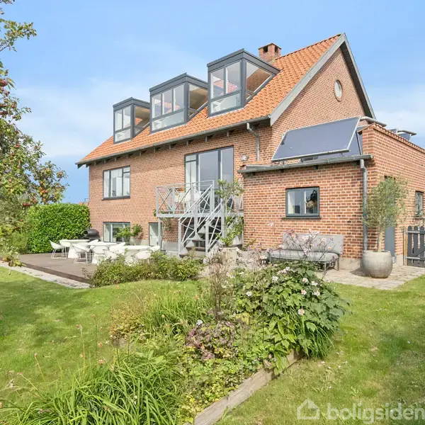 En rød murstensvilla med store vinduer har en solterrasse og en velplejet have med buske og træer, omgivet af en grøn græsplæne.